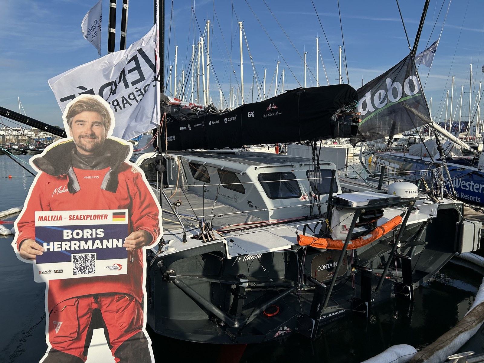 The Malizia – Seaexplorer IMOCA racing yacht moored stern-first in the race harbour, with a life-size cutout of skipper Boris Herrmann holding a sign on the pontoon